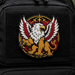 May include: A circular embroidered patch featuring a mythical griffin with a white eagle head, golden lion body, and red-tipped wings. The creature is centered within a yellow ring against a red background, all attached to a black fabric surface.