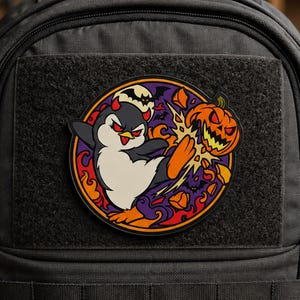 Puede incluir: Un parche circular sujeto a una mochila negra que muestra un pingüino de dibujos animados con cuernos de diablo rojos pateando una calabaza de Halloween. El fondo incluye una luna llena, murciélagos y llamas naranjas en un borde circular morado.