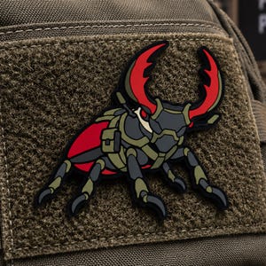 May include: A tactical morale patch featuring a stylized stag beetle illustration. The beetle is depicted with large red mandibles, a dark grey body, and olive green tactical gear straps. The design is rendered in a flat, graphic style.