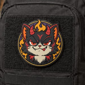Puede incluir: Un parche circular que muestra un gato de dibujos animados con cuernos rojos y una expresión traviesa. El gato tiene pelaje negro con detalles en rojo, dientes afilados y una pequeña lengua fuera sobre un fondo de llamas de color naranja.