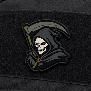May include: A black tactical morale patch featuring a grim reaper character with a white skull, red eyes, and a dark grey hooded cloak. The figure holds a scythe with a tan handle and a grey blade. A small red circular accent is visible on the cloak.