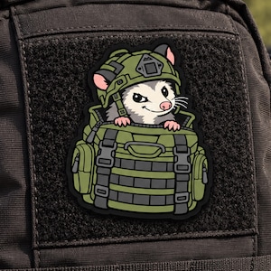 May include: A tactical morale patch featuring a cartoon opossum wearing a green military helmet and peeking out from inside a green tactical backpack with black straps and webbing, set against a black hook-and-loop fastener backing.