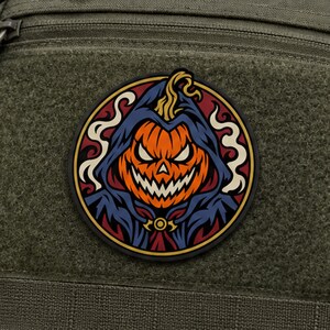 Puede incluir: Un parche circular que presenta una calabaza de Halloween naranja estilizada con una expresión amenazante, vistiendo una capa con capucha azul oscuro. El diseño incluye remolinos de humo color crema sobre un fondo rojo intenso, bordeado en negro.