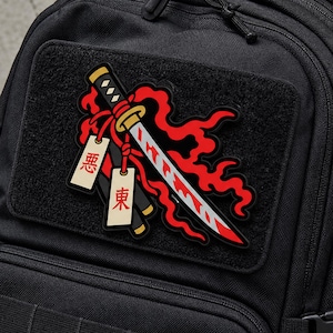 May include: A rectangular black tactical patch featuring a graphic of a katana sword surrounded by red flames. Two off-white rectangular tags with red Japanese characters hang from the sword hilt. The patch is attached to a black fabric backpack.