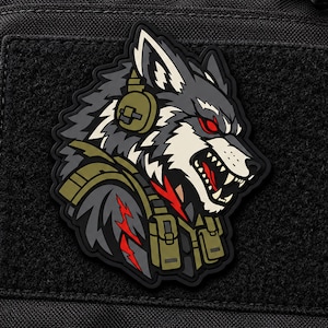 Puede incluir: Un parche táctico que muestra una cabeza de lobo estilizada con ojos rojos, vistiendo equipo táctico verde oliva y auriculares. El lobo tiene pelaje gris, negro y blanco, con dientes afilados al descubierto. El diseño incluye rayos rojos en la zona del hombro.