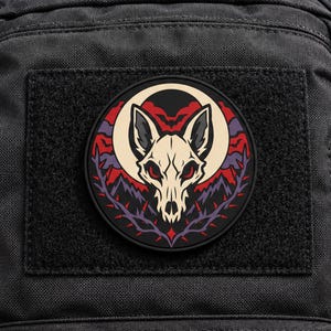 Pode incluir: Um emblema circular com um crânio de animal bege centrado contra uma grande lua pálida. O fundo inclui nuvens vermelhas, silhuetas de pequenos morcegos, montanhas roxas escuras e uma borda de vinhas espinhosas em roxo escuro e vermelho.