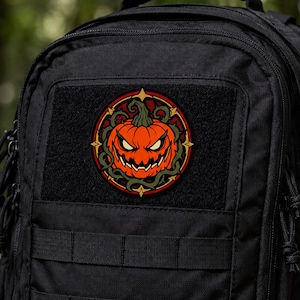 Puede incluir: Un parche circular que presenta una calabaza naranja con una expresión amenazante, rodeada de enredaderas espinosas de color verde oscuro dentro de un borde rojo y dorado, sujeto al frente de una mochila táctica negra con panel de velcro.