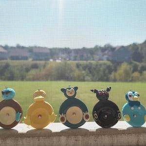 May include: Five circular ornaments featuring stylized character designs. From left to right: a blue and brown figure with sunglasses, a yellow duck-like character, a dark green and cream creature, a black figure with red eyes, and a blue and white bird.