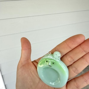 May include: A pale green, circular glass pendant featuring a smooth, glossy finish. The pendant displays a stylized face with two small black dots for eyes and a thin line for a mouth. A tiny, round, light green figure sits atop the main piece.