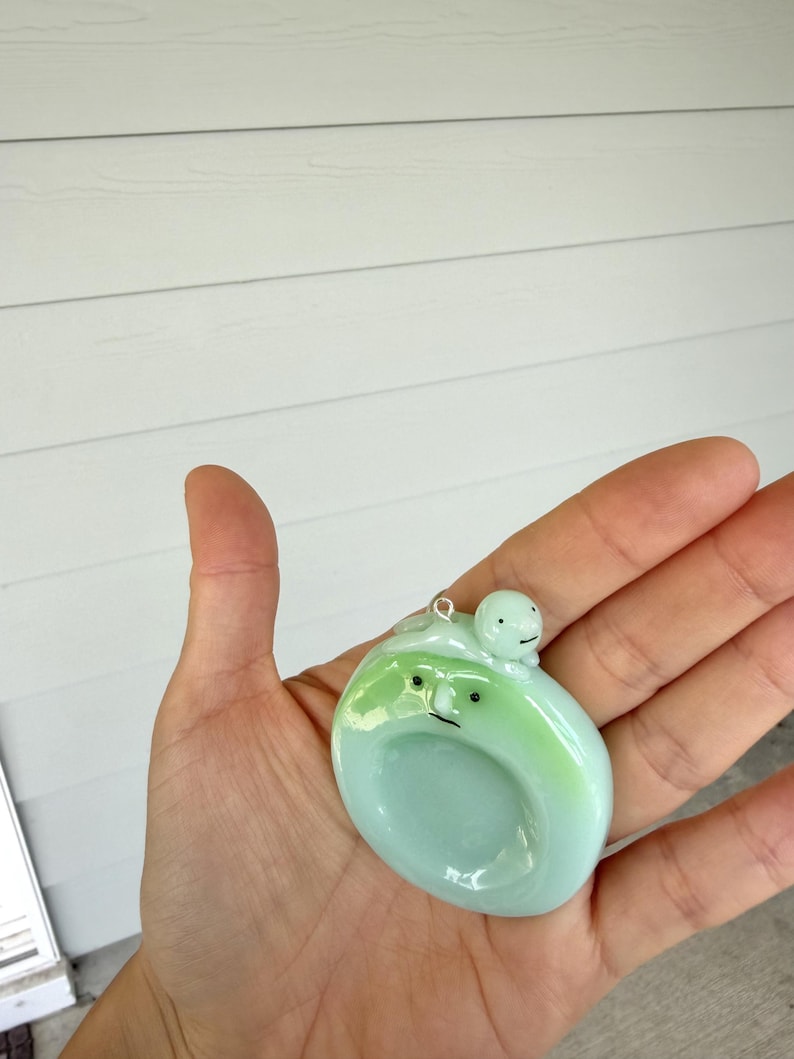 May include: A pale green, circular glass pendant featuring a smooth, glossy finish. The pendant displays a stylized face with two small black dots for eyes and a thin line for a mouth. A tiny, round, light green figure sits atop the main piece.