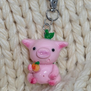 May include: A pink pig figurine keychain featuring a small green leaf on its head and holding a miniature orange peach in its hand. The pig has black eyes and a silver-toned metal lobster clasp attached to a circular ring at the top of its head.