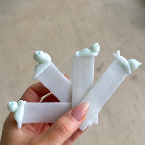 May include: Four white rectangular bookmarks, each topped with a small, pale green glass cat figurine. The cats are depicted in various resting positions, including lying down and sitting, with subtle green accents on their faces and ears.