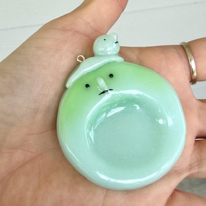 May include: A round, light green ceramic pendant with a glossy finish. The pendant features a small, sculpted figure on top and a face with two black dots for eyes and a thin black line for a mouth. A small metal jump ring is attached to the top.