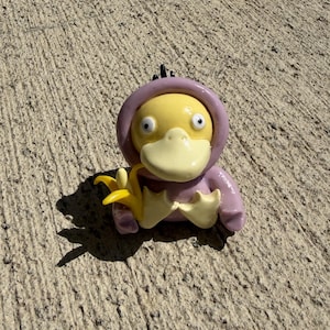 May include: A small, glossy figurine of a yellow duck-like creature with a wide beak and vacant eyes, wearing a smooth lavender-colored hooded garment. The figure is holding a small, bright yellow banana in its webbed hands, with black hair tufts.
