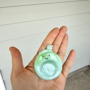 May include: A round, light green glass pendant featuring a small, sculpted face with two black dot eyes and a thin line mouth. A tiny, smooth, light green bird-like figure sits on the top edge of the pendant, which includes a small metal loop.