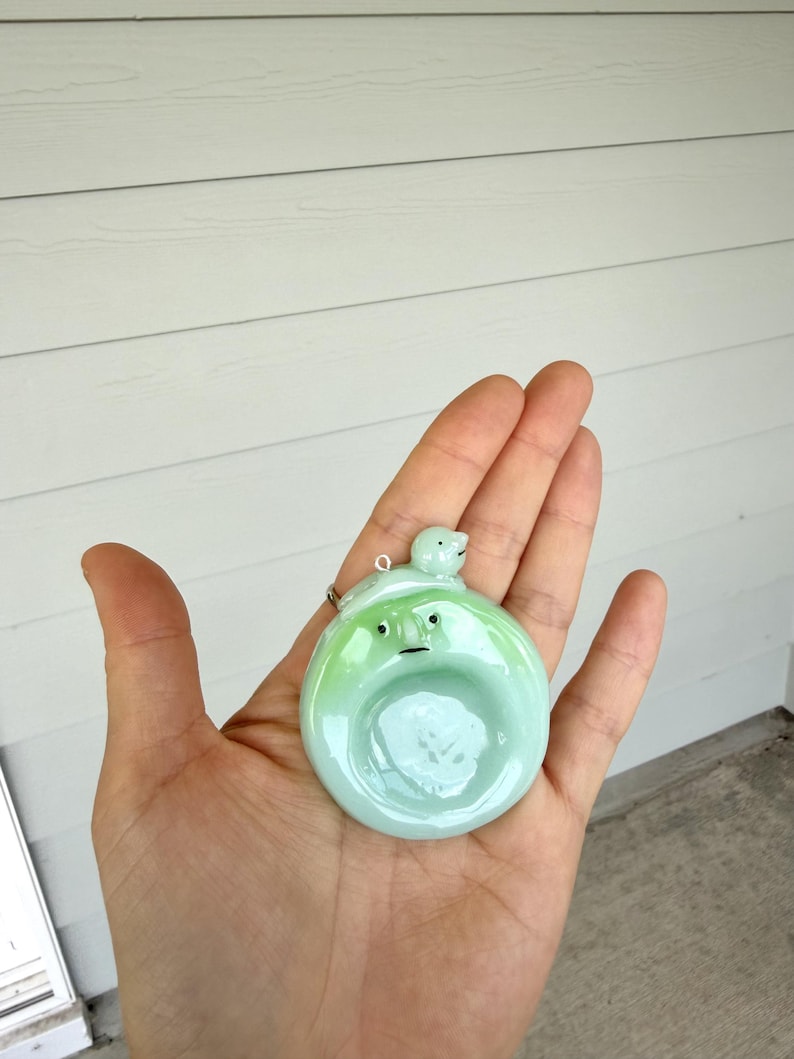 May include: A round, light green glass pendant featuring a small, sculpted face with two black dot eyes and a thin line mouth. A tiny, smooth, light green bird-like figure sits on the top edge of the pendant, which includes a small metal loop.