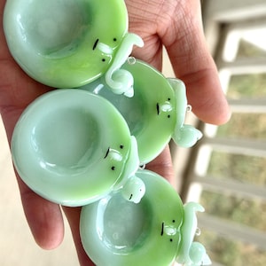 May include: Three circular, light green glass charms, each featuring a small, sculpted white figure attached to the rim. The glass has a smooth, glossy finish with subtle color variations. Each charm includes a small metal loop for hanging.