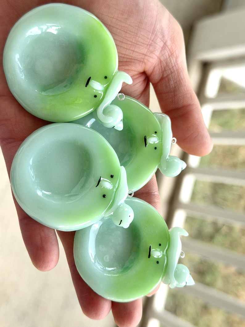May include: Three circular, light green glass charms, each featuring a small, sculpted white figure attached to the rim. The glass has a smooth, glossy finish with subtle color variations. Each charm includes a small metal loop for hanging.
