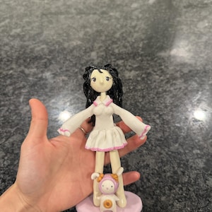 May include: A handcrafted clay figurine featuring a character with long black hair, wearing a white dress with pink trim, standing on a round light purple base. A small, white, rabbit-like creature with a pink hood and a brown donut detail stands at the feet.