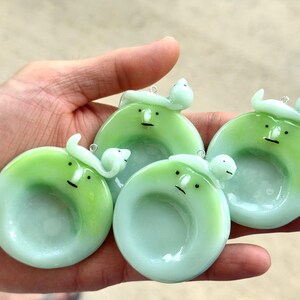 May include: Three circular, light green glass pendants featuring a stylized face with a small nose and a flat line for a mouth. Each pendant has a small, curved, light green glass figure attached to the top, complete with a metal jump ring.