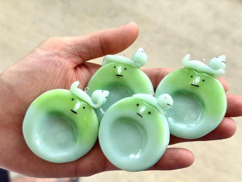 May include: Three circular, light green glass pendants featuring a stylized face with a small nose and a flat line for a mouth. Each pendant has a small, curved, light green glass figure attached to the top, complete with a metal jump ring.