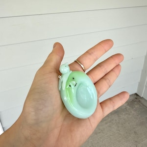 May include: A pale green, oval-shaped glass pendant featuring a smooth, glossy finish with a small, sculpted figure resting on top. The pendant has a subtle gradient of light green and includes a small metal loop for attachment to a necklace chain.