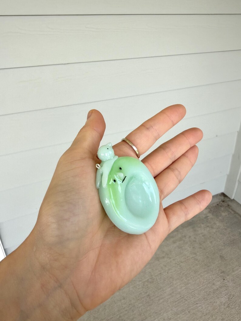 May include: A pale green, oval-shaped glass pendant featuring a smooth, glossy finish with a small, sculpted figure resting on top. The pendant has a subtle gradient of light green and includes a small metal loop for attachment to a necklace chain.