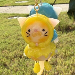 May include: A resin keychain featuring a cat figure wearing a bright yellow hooded raincoat with a bow at the neck, dangling from a gold-toned metal ring. The cat has black eyes, whiskers, and a pink nose, with a second blue figure visible behind.