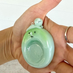 May include: A smooth, oval-shaped pendant made of light green glass with a glossy finish. The pendant features a sculpted face with two small black dots for eyes and a thin line for a mouth. A tiny, round, light green creature sits on top.