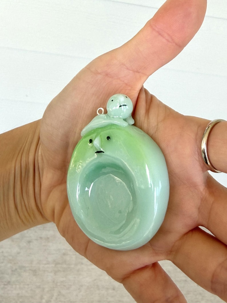 May include: A smooth, oval-shaped pendant made of light green glass with a glossy finish. The pendant features a sculpted face with two small black dots for eyes and a thin line for a mouth. A tiny, round, light green creature sits on top.