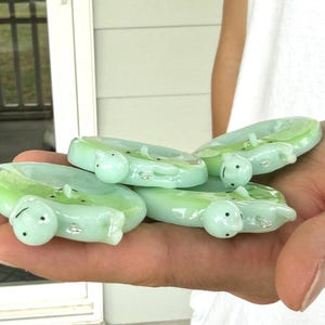May include: A collection of three pale green glass rings, each featuring a small, sculpted turtle figure attached to the band. The turtles have smooth, rounded shells and tiny black dots for eyes, with a glossy, translucent finish throughout.