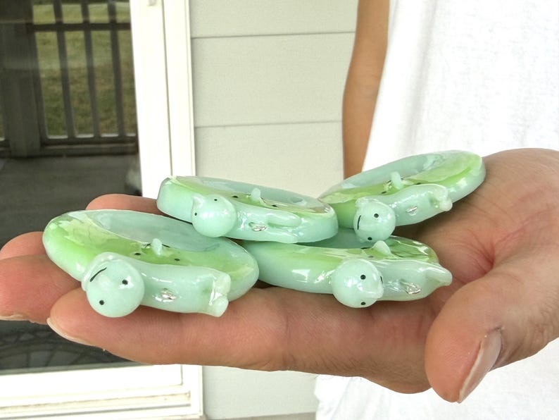 May include: A collection of three pale green glass rings, each featuring a small, sculpted turtle figure attached to the band. The turtles have smooth, rounded shells and tiny black dots for eyes, with a glossy, translucent finish throughout.