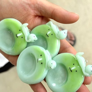 May include: Four circular glass pendants in shades of light green and mint, each featuring a small, sculpted creature with two black dot eyes and a simple line mouth. Each pendant includes a small metal jump ring for attachment to a necklace.