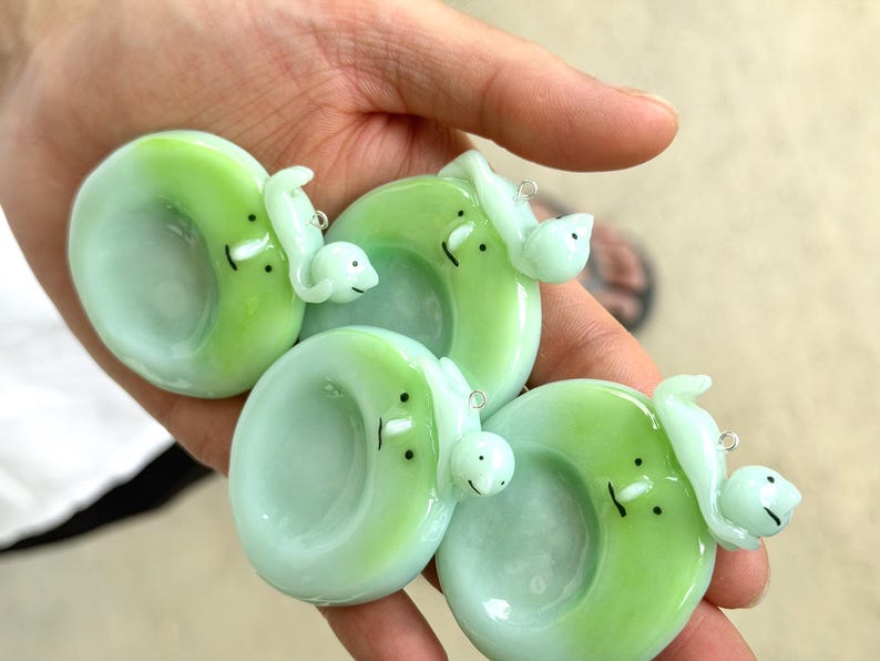 May include: Four circular glass pendants in shades of light green and mint, each featuring a small, sculpted creature with two black dot eyes and a simple line mouth. Each pendant includes a small metal jump ring for attachment to a necklace.
