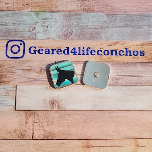 May include: Two small, square, metal concho buttons. One button features a turquoise and white design with a black silhouette of a donkey's head. The other button is silver with a smooth surface. The buttons are on a wooden background.  The text "Geared4lifeconchos" is visible in the image.