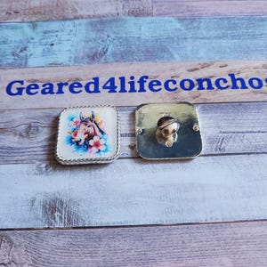 May include: A silver metal concho with a square shape and a horse head design. The concho has a white background with pink flowers and a brown horse head. The concho is surrounded by a silver border.