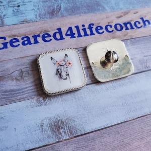 May include: A silver square-shaped pin with a white background and a black and white illustration of a donkey wearing a flower crown. The pin has a silver border and a silver back with a screw post.