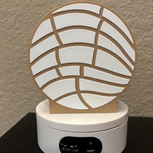 May include: A circular tan and white concha-shaped object mounted on a white rotating display base. The base features a black control panel with the text CW/OFF/CCW and a DC-5V port. The top piece has a textured white grid pattern on a tan background.