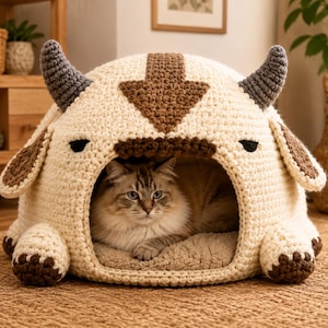 May include: A cream-colored crocheted pet bed shaped like a creature with two dark gray horns, brown ear flaps, and a large brown arrow design on top. A fluffy, long-haired cat with brown and white fur is resting inside the open, cave-like structure.