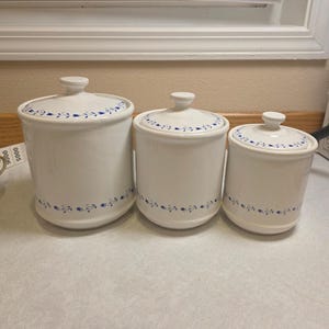 Op de afbeelding: Een set van drie witte keramische voorraadpotten met bijpassende deksels, elk voorzien van een delicaat blauw bloemenrankpatroon rond de basis en de dekselrand. De potten zijn gerangschikt in afnemende grootte van links naar rechts.