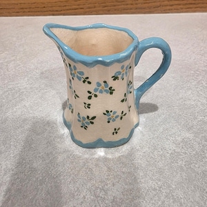 Puede incluir: Una jarrita de cerámica para crema con cuerpo de color crema, decorada con un delicado patrón de flores azules y hojas verdes. La jarra presenta un borde festoneado y un asa pintada en azul claro, con un acabado de esmalte craquelado visible.