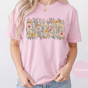 Könnte beinhalten: Ein hellrosa T-Shirt mit kurzen Ärmeln und dem Wort BLESSED in großen Blockbuchstaben auf der Brust. Die Buchstaben sind mit einem detaillierten Blumenmuster gefüllt, das aus kleinen Blüten, Blättern und Stängeln in Rosa-, Gelb- und Grüntönen besteht.