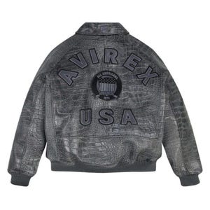 May include: A charcoal grey leather bomber jacket with a textured crocodile skin pattern. The back features the text AVIREX and USA in dark grey lettering, along with a circular patch displaying the text ALL AMERICANS ESTABLISHED 1975.