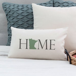 May include: White throw pillow with the word "HOME" in black text. The letter "O" is replaced with the outline of the state of Minnesota in green.