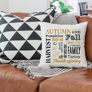 May include: A white square pillow with the words "Autumn", "Harvest", "Fall", and "Thanksgiving" in black and gold lettering. The pillow is on a brown leather couch with two other pillows, one black and white geometric and one striped.