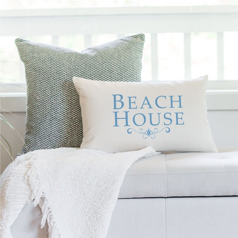 May include: A white throw pillow with the text "BEACH HOUSE" in blue lettering and a decorative flourish. The pillow is sitting on a white sofa with a green and white patterned pillow behind it.