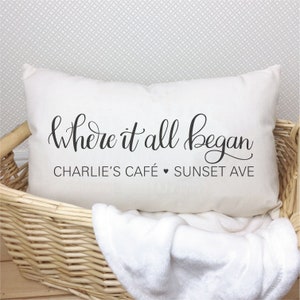 Puede incluir: Coj&iacute;n rectangular color crema con el texto "Where it all began" en escritura negra. Debajo, se lee "Charlie's Caf&eacute;" y "Sunset Ave". El coj&iacute;n est&aacute; en una cesta de mimbre con una manta blanca.