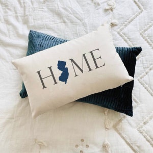 May include: A white pillow with the word "HOME" printed in gray with a blue outline of the state of New Jersey. The pillow is sitting on top of a blue velvet pillow.