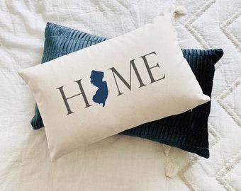 Puede incluir: Una almohada blanca con la palabra "HOME" impresa en gris con un contorno azul del estado de Nueva Jersey. La almohada está encima de una almohada de terciopelo azul.