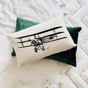 May include: A white cushion with a black and white illustration of a vintage biplane. The cushion is sitting on top of a dark green cushion.